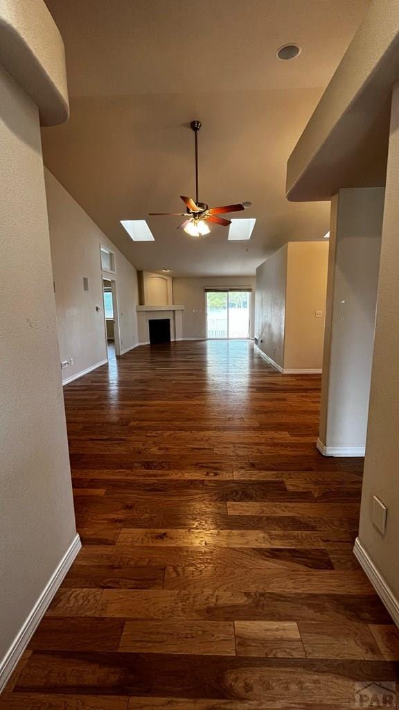 unfurnished living room featuring vaulted ceiling with skylight, dark wood-style flooring, ceiling fan, and baseboards