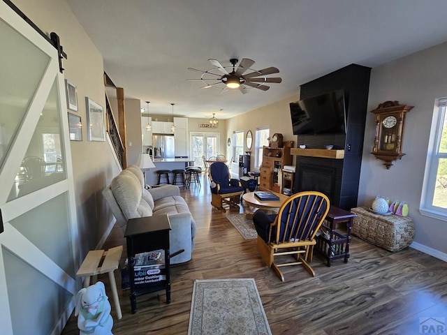 living area with ceiling fan with notable chandelier, dark wood finished floors, a barn door, a fireplace, and baseboards