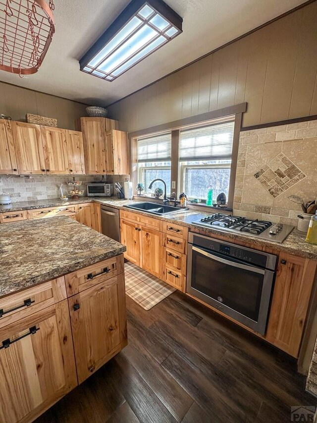 kitchen with dark wood-style flooring, backsplash, appliances with stainless steel finishes, a sink, and dark stone counters