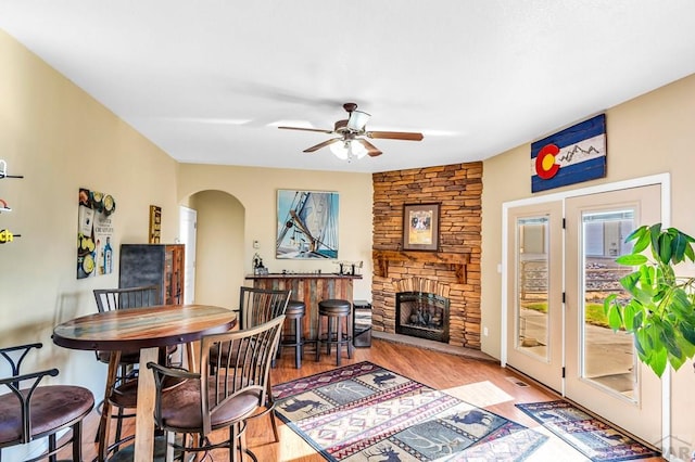 dining space featuring a stone fireplace, wood finished floors, arched walkways, and ceiling fan
