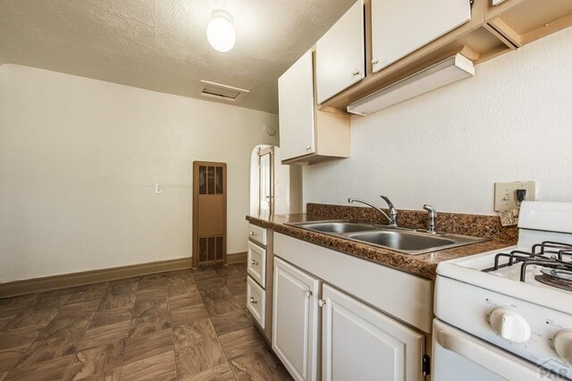 kitchen featuring white gas range, dark countertops, white cabinets, a sink, and baseboards