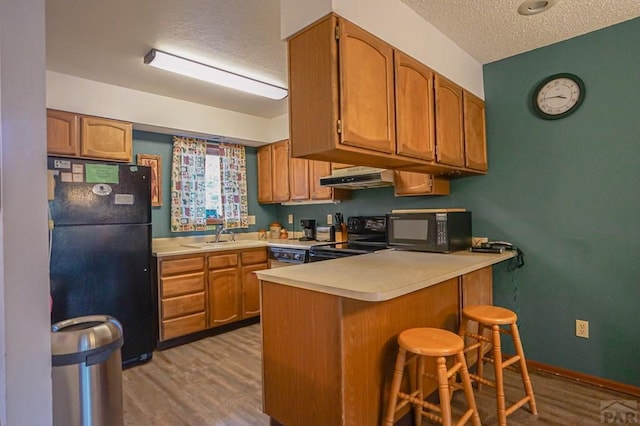 kitchen with under cabinet range hood, a peninsula, light countertops, black appliances, and a kitchen bar