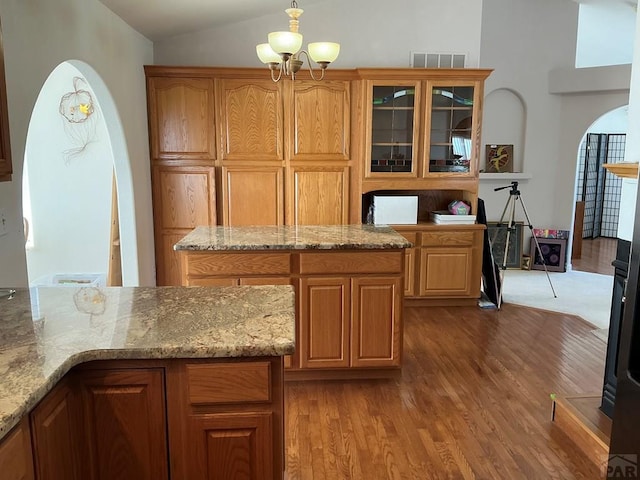 kitchen with arched walkways, vaulted ceiling, light stone countertops, light wood finished floors, and glass insert cabinets