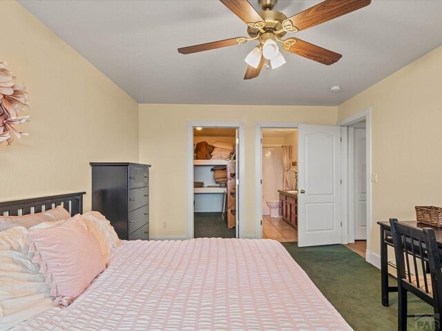 bedroom featuring a walk in closet, dark carpet, connected bathroom, baseboards, and ceiling fan