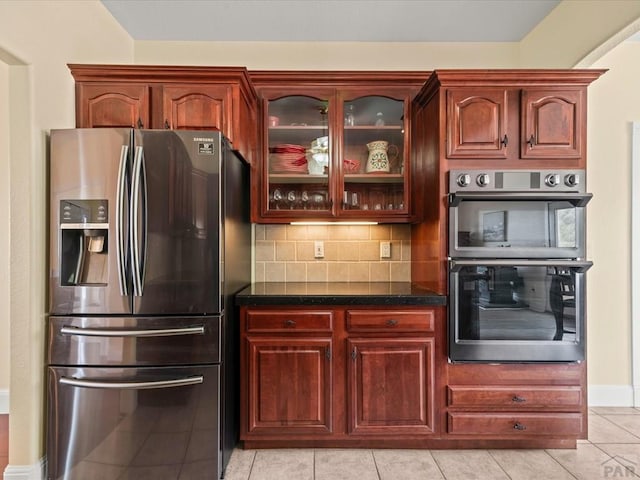 kitchen with dark stone countertops, backsplash, appliances with stainless steel finishes, light tile patterned floors, and glass insert cabinets