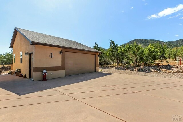 view of side of property with a mountain view, an outdoor structure, a detached garage, roof with shingles, and stucco siding