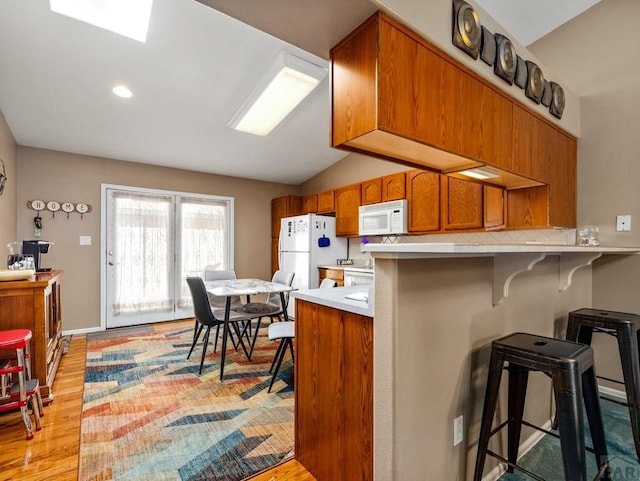 kitchen featuring a breakfast bar, brown cabinets, vaulted ceiling, light wood-type flooring, and white appliances