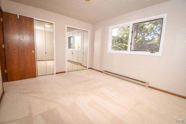 unfurnished bedroom featuring a textured ceiling, carpet floors, baseboard heating, multiple windows, and two closets