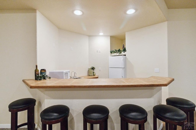 kitchen with a breakfast bar, tile counters, recessed lighting, freestanding refrigerator, and a sink