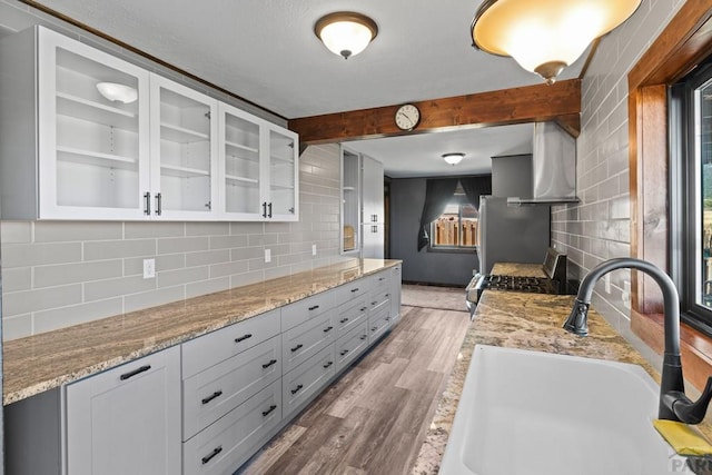 kitchen featuring light wood finished floors, appliances with stainless steel finishes, glass insert cabinets, wall chimney range hood, and a sink