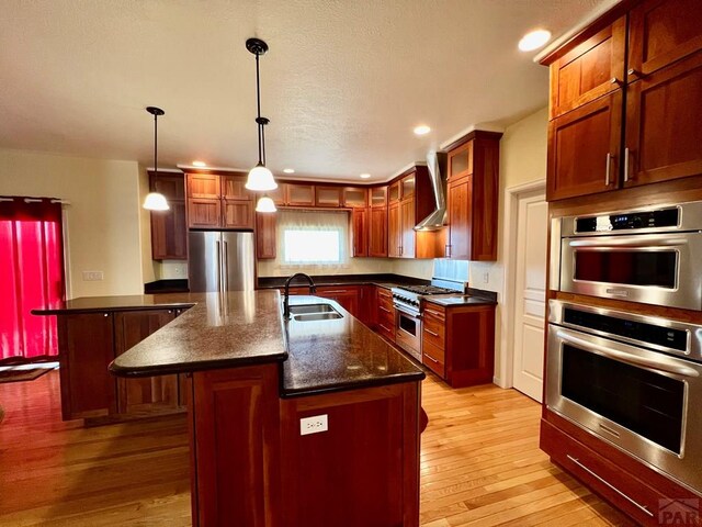 kitchen with pendant lighting, a center island with sink, stainless steel appliances, a sink, and wall chimney exhaust hood