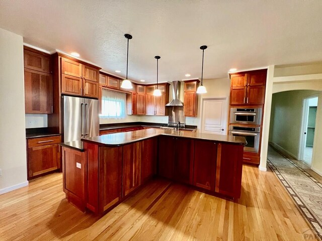 kitchen featuring an island with sink, wall chimney exhaust hood, appliances with stainless steel finishes, and hanging light fixtures