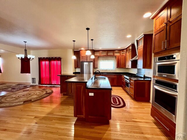 kitchen with stainless steel appliances, dark countertops, a kitchen island with sink, and decorative light fixtures