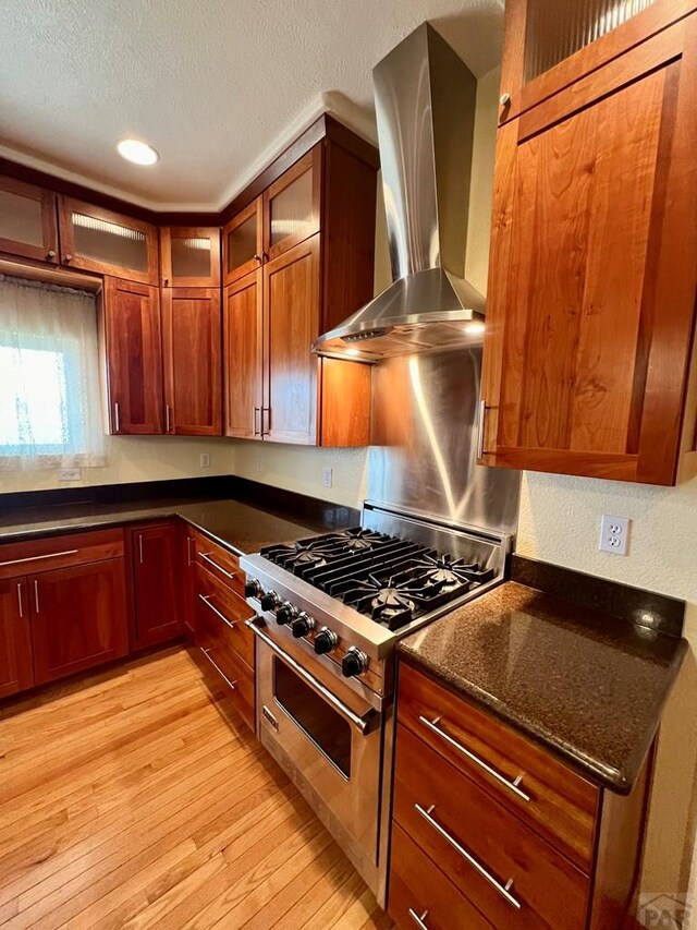 kitchen with light wood finished floors, glass insert cabinets, a textured ceiling, stainless steel stove, and wall chimney range hood