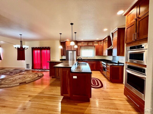 kitchen featuring dark countertops, a center island with sink, appliances with stainless steel finishes, and hanging light fixtures