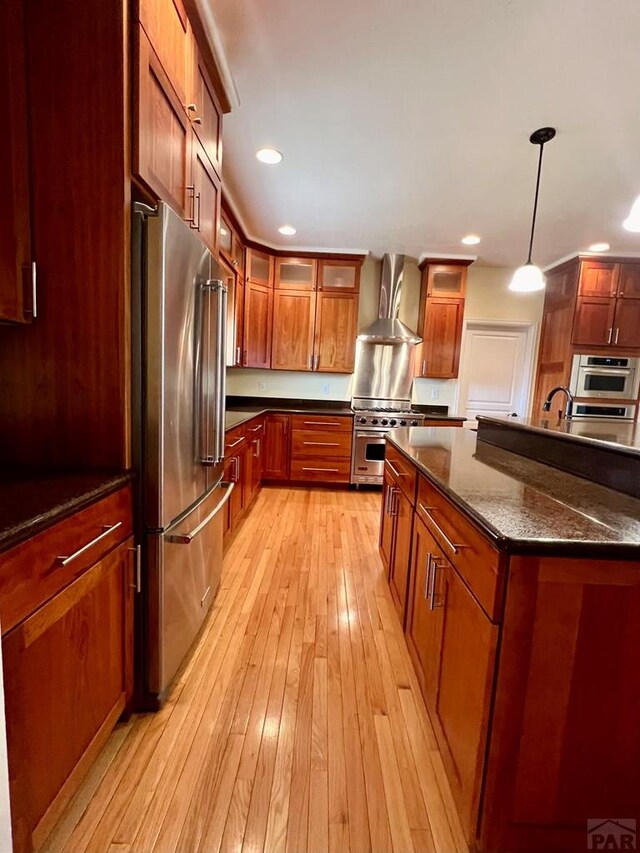 kitchen with hanging light fixtures, dark stone counters, wall chimney exhaust hood, high end appliances, and glass insert cabinets