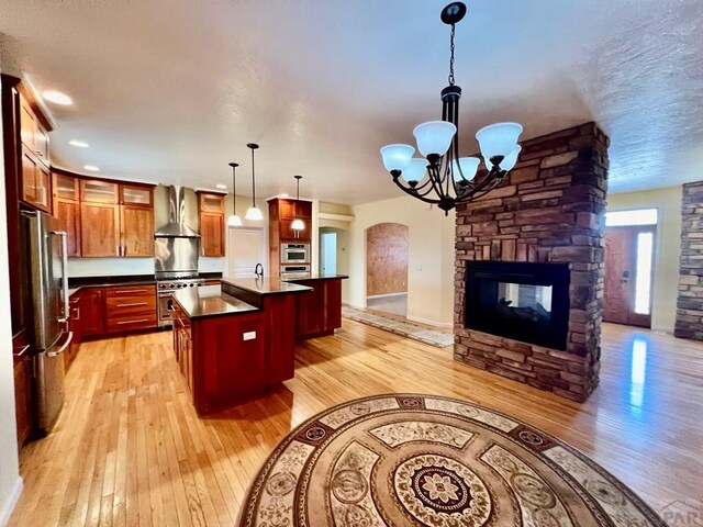 kitchen featuring decorative light fixtures, a center island with sink, stainless steel appliances, open floor plan, and wall chimney range hood