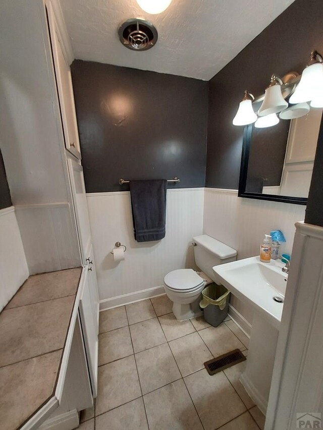 bathroom with a textured ceiling, tile patterned flooring, toilet, visible vents, and wainscoting