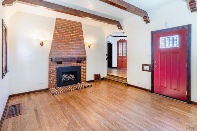 unfurnished living room featuring light wood-style flooring, visible vents, baseboards, a brick fireplace, and beamed ceiling