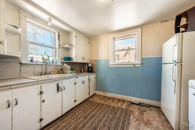 kitchen with open shelves, freestanding refrigerator, wainscoting, white cabinetry, and a sink