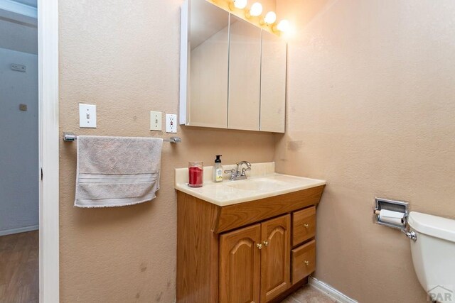 half bath with toilet, a textured wall, vanity, and baseboards