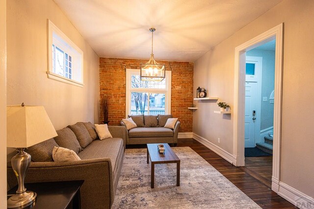 living room with a chandelier, brick wall, dark wood-type flooring, baseboards, and stairway