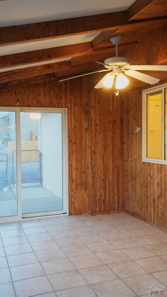 empty room featuring vaulted ceiling with beams, light tile patterned floors, wooden walls, and a ceiling fan