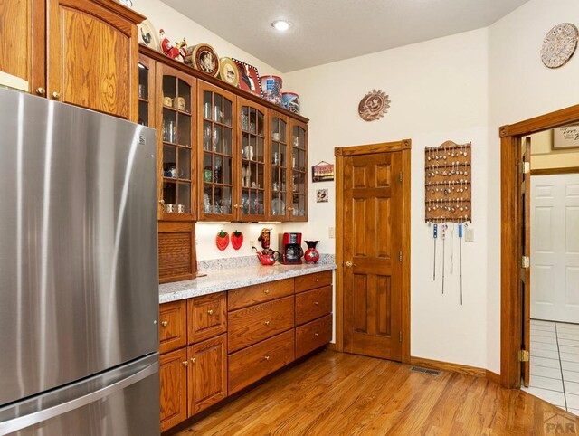 kitchen with glass insert cabinets, freestanding refrigerator, light wood-type flooring, and light stone countertops