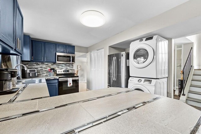 kitchen with blue cabinetry, light countertops, appliances with stainless steel finishes, a sink, and stacked washing maching and dryer