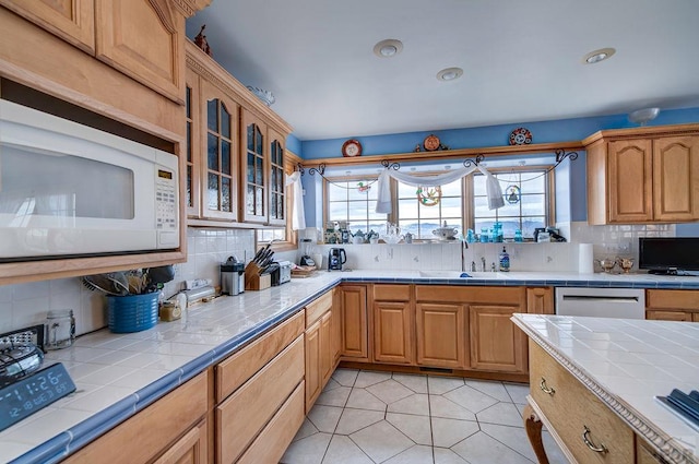 kitchen with tile countertops, stainless steel dishwasher, white microwave, glass insert cabinets, and a sink