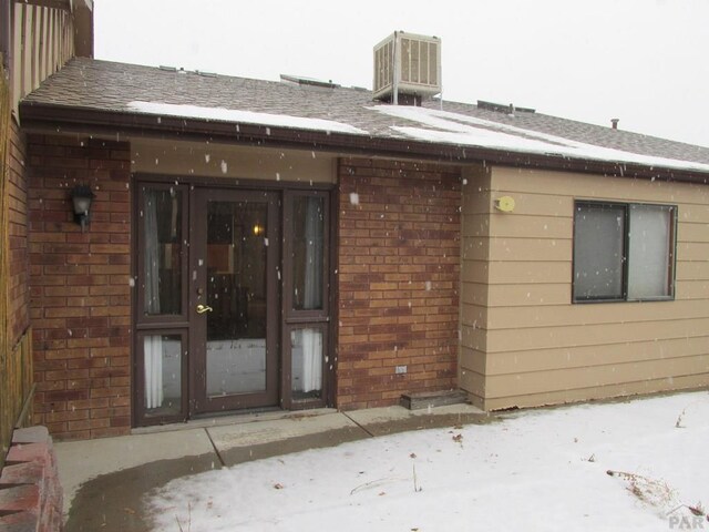 snow covered property entrance with central air condition unit, a shingled roof, and brick siding