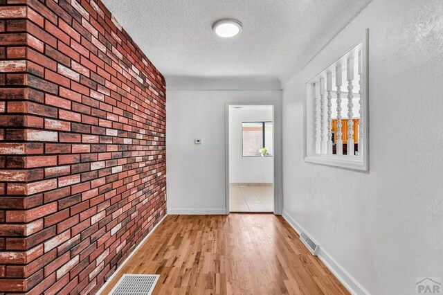 hallway with visible vents, a textured ceiling, brick wall, light wood-type flooring, and baseboards