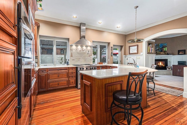 kitchen with a kitchen island with sink, stainless steel appliances, light countertops, wall chimney range hood, and a kitchen bar