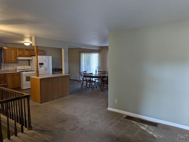 kitchen featuring under cabinet range hood, white appliances, light countertops, brown cabinetry, and dark carpet