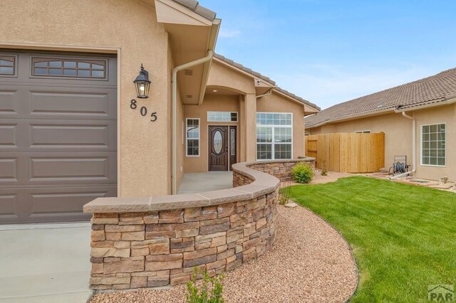 view of exterior entry featuring a garage, fence, a lawn, and stucco siding